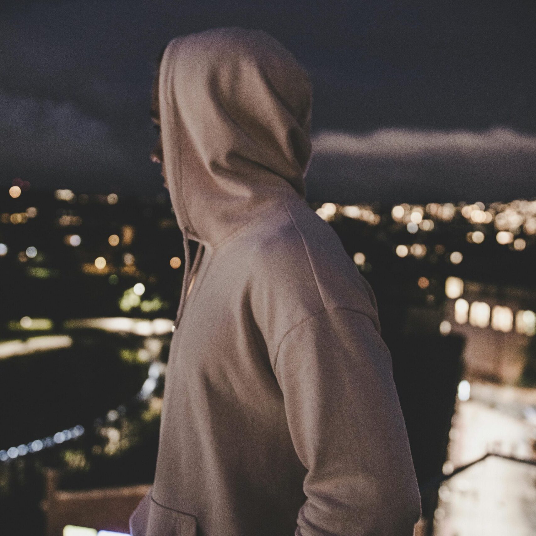 A person in a hoodie overlooks a blurry cityscape at night, evoking mystery and solitude.